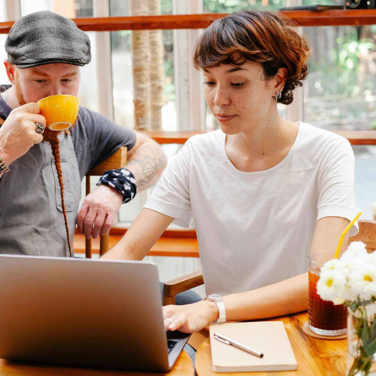 A woman is focused on a laptop, while a man sits beside her sipping coffee from a yellow mug. The wooden table features a notebook, pen, and a glass of iced drink with a yellow straw, accompanied by a small bouquet of white flowers.